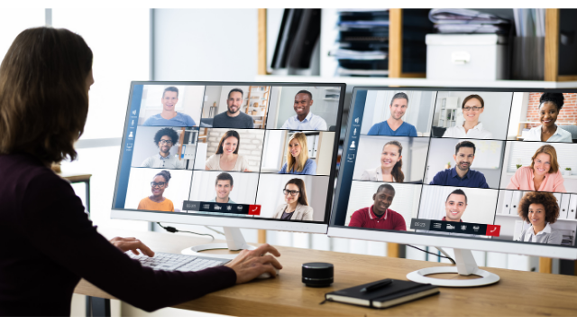 Woman with 2 large monitors on her desk (and.a journal) with 9 squares of diverse faces on each monitor