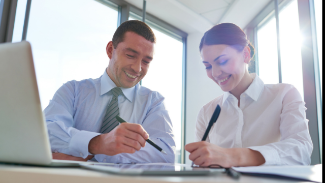 Business meeting in conference room with a computer open, and 2 people with pens smiling and talking