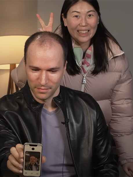 A man in a black leather jacket is holding a smartphone, showing a photo of himself on the screen. Behind him, a woman wearing a puffy jacket and a scarf is smiling widely and playfully making bunny ears with her fingers above the man's head. They are both indoors, with a lamp in the background.