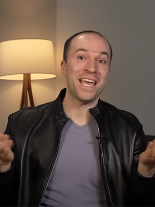 A man in a black leather jacket is smiling and gesturing with his hands while sitting indoors. He has a cheerful expression and is wearing a light purple shirt underneath the jacket. In the background, there is a lamp providing warm lighting.