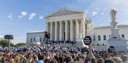 A large crowd gathered in front of the U.S. Supreme Court building, with its iconic columns and neoclassical architecture in the background. Many people in the crowd are holding signs, including one prominently displayed in the foreground that reads "Stop Election Fraud." Some individuals, possibly law enforcement or court officials, appear to be dressed in dark uniforms or robes. The atmosphere suggests a protest or rally, with a mix of cameras and raised hands capturing the event. Scaffolding is visible on part of the building, indicating construction or maintenance work. The sky is clear with a few scattered clouds.