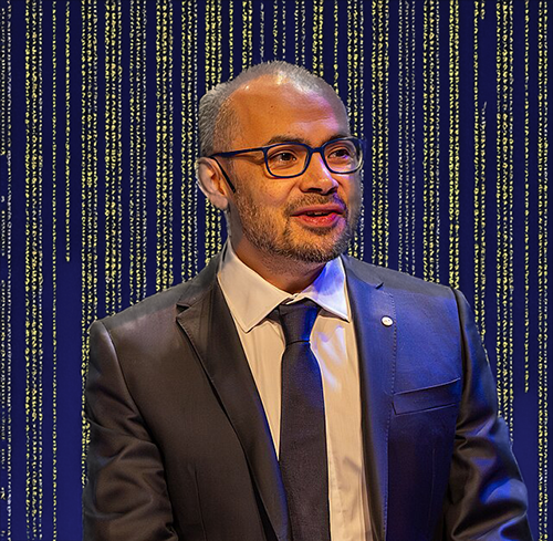 A professional portrait of a man with a shaved head and glasses, dressed in a black suit, white shirt, and dark tie. He is smiling slightly and appears to be speaking or presenting. The background has been digitally edited to feature cascading golden lines resembling digital code, set against a deep blue backdrop. The effect creates a futuristic, high-tech aesthetic, symbolizing innovation and technology. The man exudes confidence and professionalism in this formal setting.